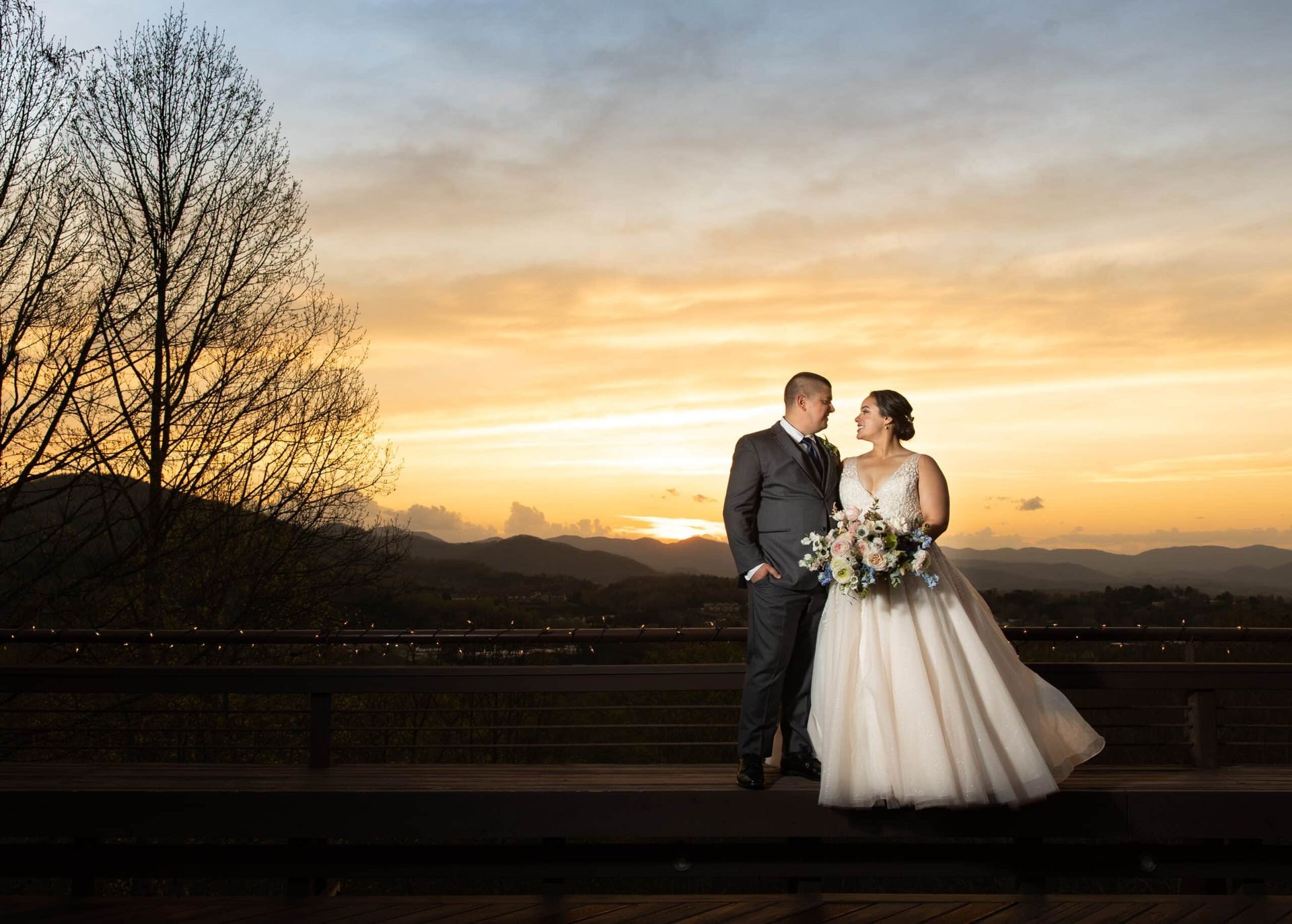 Bride and groom standing on a deck at sunset with mountains in the background.