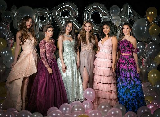 Six young women in elegant dresses standing together with balloons and 'PROM' letters in the background.